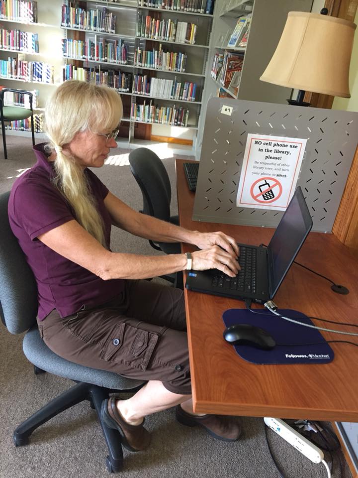 Gretchen sitting at a library computer workstation typing on a laptop.